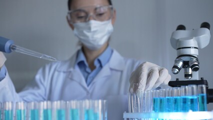 Scientist woman, wearing a lab coat, mask, blue gloves and safety glasses, is using a micropipette...