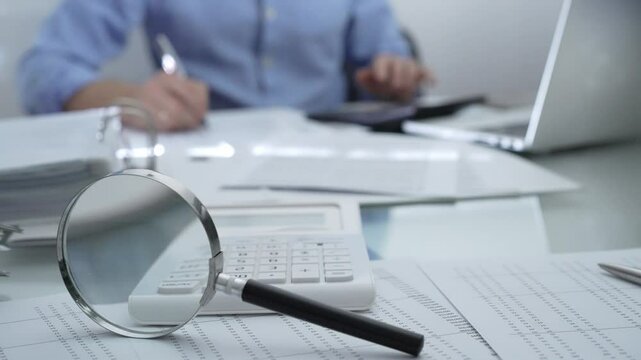 Magnifying glass, calculator, pen and documents are lying on accountant's desk. Businessman wearing blue shirt is working with documents on the background. Business people, audit concept