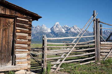 Fototapeta premium Rustic Cabin with Wooden Fence and Grand Tetons Mountain Range Under Clear Blue Sky..