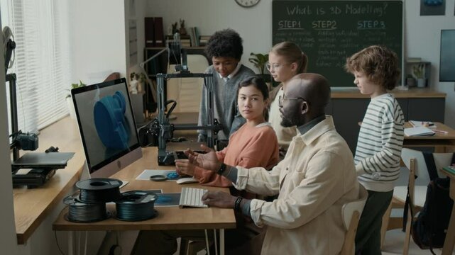 African American male teacher sitting at computer table and explaining 3D modelling to young students, surrounding him in modern elementary school