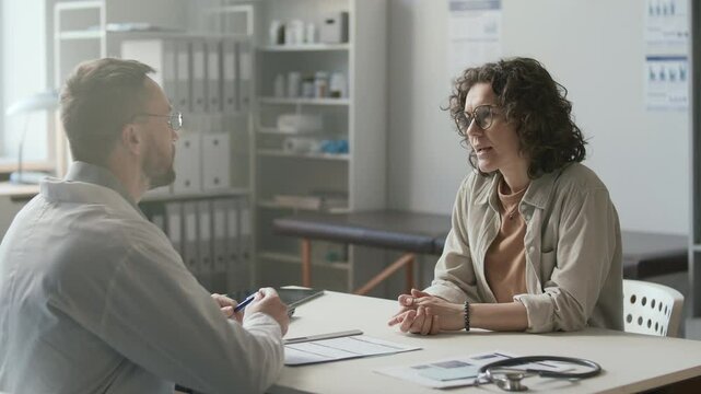 Female patient explaining health concerns and having discussion with male physician in white coat, both sitting at desk in sunlit medical office