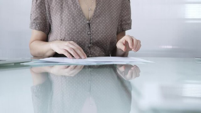 Businesswoman or accountant in brawn dotted blouse is going through paperwork while holding documents at a glass desk in a well lit, contemporary office setting. Business people concept