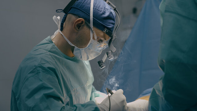 Close up doctor man physician practitioner surgeon medic in protective medical mask perform surgical operation microscope plastic surgery cautery modern medical treatment medicine hospital health care