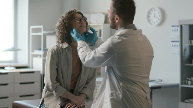 Doctor in white coat and sterile gloves examining neck of female patient and having discussion with her during medical checkup in clinic
