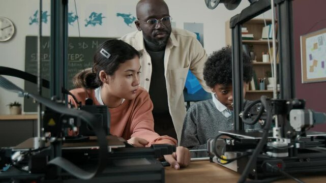 African American male teacher guiding two students, using 3D printers during 3Dm modeling class in modern school