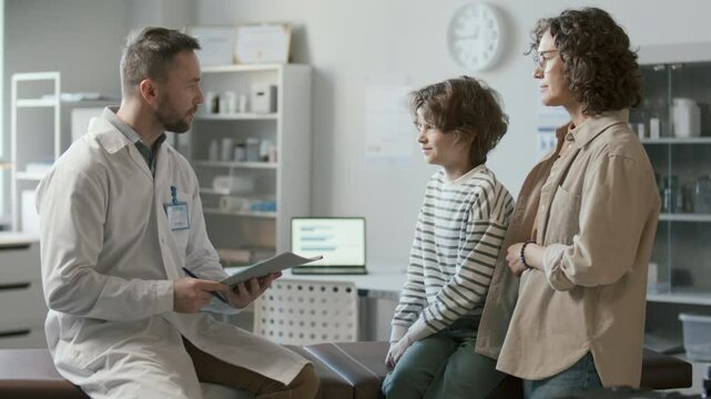 Doctor in white coat reviewing document on clipboard, listening to mother and kid telling about health concerns and giving advice during consultation in medical office