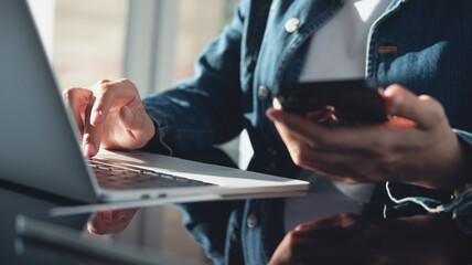 Closeup, business woman sitting at table using mobile smrt phone during working on laptop computer at home office. Young asian woman freelancer  surfing the internet, online working from home