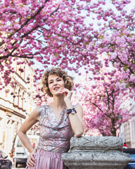 Elegant beautiful woman posing in decent outfit with cherry blossom trees in background. She look dreamy and very confident. Looking away from camera