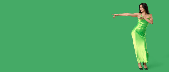 Beautiful young woman in stylish green dress dancing and pointing at something on color background