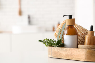 Bottles of cosmetic products, green leaves and bath supplies in wooden tray on table in bathroom, closeup