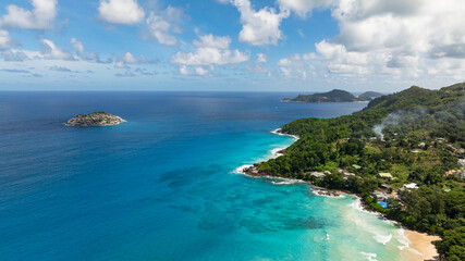 A tropical coastline with turquoise waters, a small offshore island, and green hills in the background. Seychelles, Mahe.