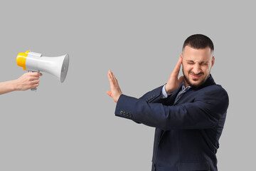 Young businessman suffering from loud noise and hand with megaphone on grey background