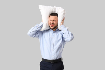 Young businessman with pillow suffering from loud noise on grey background