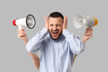 Young businessman suffering from loud noise and hands with megaphones on grey background