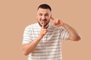 Young man suffering from loud noise and showing silence gesture on beige background