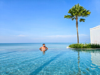 Obraz premium Enjoying the serenity of Krabi Thailand, a man relaxes in the infinity pool while surrounded by stunning blue waters and swaying palm trees under a clear sky.