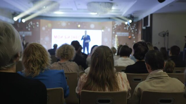 Large crowd of people are sitting in chairs in a room, watching a speaker. The speaker is on a stage in front of a large screen. Scene is one of anticipation and excitement