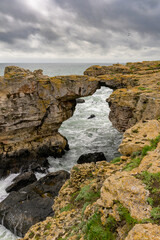 A wide-angle shot of high cliffs, rocky arc, and the sea with clouds