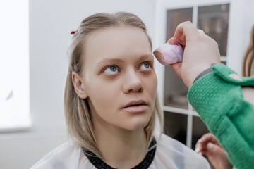 Makeup artist applying foundation, highlighter, powder with a brush to blonde's face in a beauty...