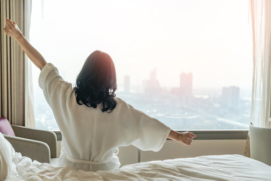 Hotel relaxation on lazy day with Asian woman take it easy, waking up from good sleep on bed in weekend morning resting in comfort bedroom city view having happy work-life quality balance lifestyle