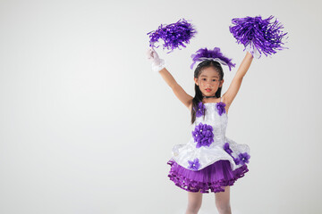 Little girl in cheerleading outfit with purple pom-poms, beaming happily in a white studio setup. A...