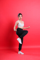 A woman in athletic attire performing a fitness pose with one leg lifted. She has black pants, white shoes and a white bra isolated on a red background