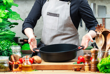 Chef holds cast iron saucepan in hands. Cozy kitchen with wooden table, kitchenware, vegetables, herbs and ingredients for cooking. Healthy eating, culinary, recipes, food blogging