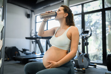 Fototapeta premium Portrait of a beautiful young pregnant woman drinking her coffee in the gym. Working out, yoga and fitness, pregnancy concept. 