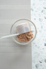 A top down view of a measuring scoop pouring protein powder into a small glass bowl.