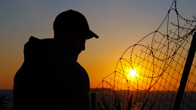 Man gazing at sunset behind barbed wire fence. Silhouette of a refugee man wearing a baseball cap looking at sunset behind a barbed wire fence, evoking themes of confinement, longing, and hope
