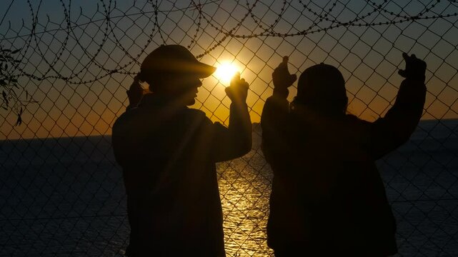 Refugee family at barbed wire at sunset. Mother and daughter hold on to the barbed wire and watch the sunset with hope for freedom and a new life.