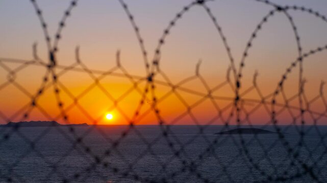 Orange sunset behind barbed wire over calm sea. Barbed wire fence dominates the foreground, obscuring a tranquil ocean sunset view, evoking longing, and the contrast between freedom and captivity