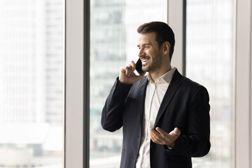 Young businessman, company boss leads formal talk by smartphone, standing near panoramic window...