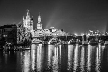 Fototapeta premium Illuminated towers and arches of Charles Bridge reflect in the tranquil Vltava River during a serene night in Prague. The city lights create a stunning nighttime ambiance.