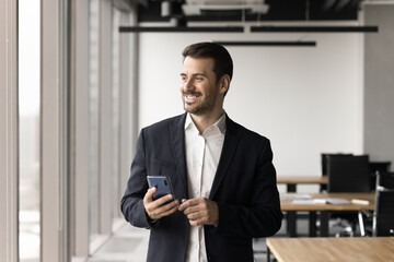 Smiling handsome businessman dressed in suit holding smart phone standing by panoramic window in modern co-working office, making business calls, chatting with client, receive good professional news