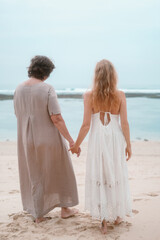 An elderly mother and her adult red haired daughter in white dresses are holding hands by the ocean. They are talking to each other and laughing. Family vacation. Mothers Day.