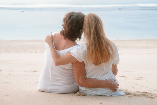 Back view of a senior mother and her young redhead daughter sitting on the beach during a family vacation. They hug and look at the ocean. Mothers Day.