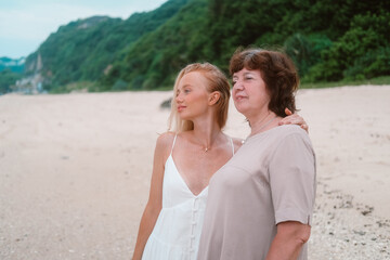 A young redhead charming woman tenderly hugs her senior mother standing on the beach near the ocean...