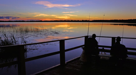 Two anglers fishing from a wooden pier at sunset by a calm lake surrounded by lush greenery and...