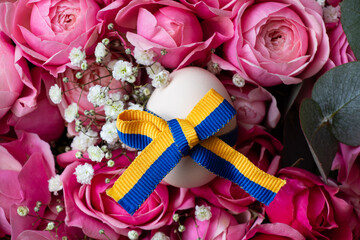 Ukrainian flag ribbon on chicken egg, egg lies on bouquet of pink roses, Easter holiday