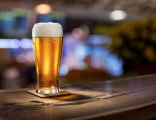 Glass of chilled beer on wooden bar tabletop and blurred brightly lit bar interior in the background.