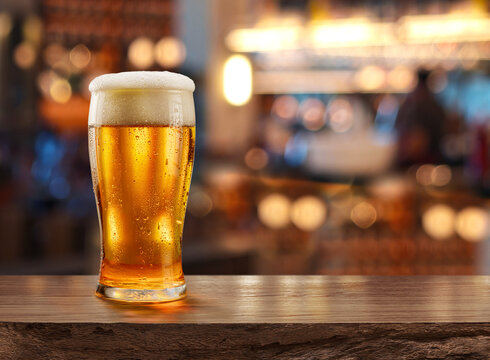 Glass of chilled beer on wooden bar tabletop and blurred brightly lit bar interior in the background.