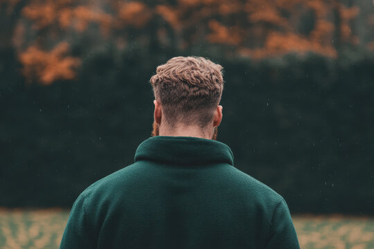 Back view of a man with curly hair wearing a dark green coat standing in an autumn park
