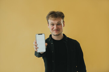 Young Man Smiling and Displaying a Smartphone Against a Yellow Background