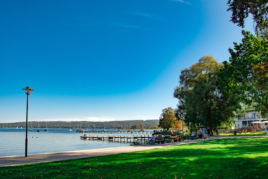 Peaceful lakeside promenade with green grass, trees, dock and clear blue sky on a sunny day in Bavaria
