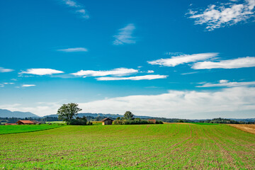 Rural landscape in Bavaria with green fields, plowed soil, farmhouse and distant mountains under blue sky

