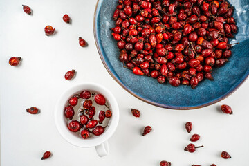 Hot rose hip herbal tea in a white cup with dried red berries on a ceramic round plate on a white wooden table. Medicinal plants. Dog Rose background. Top view.