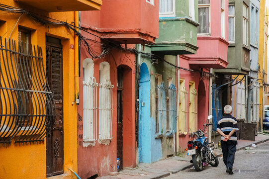 A charming, narrow street in Balat, Istanbul, featuring colorful historic houses with traditional bay windows, where an elderly man walks past a parked motorcycle, reflecting the authentic and vibrant