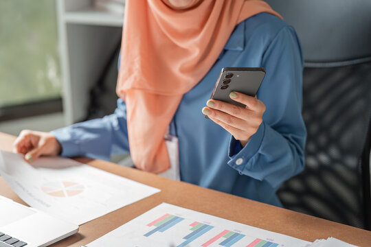 Close-up of Muslim woman in hijab using smartphone while reviewing financial reports at a desk.