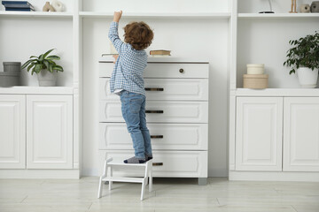 Little boy standing on step stool near chest of drawers at home